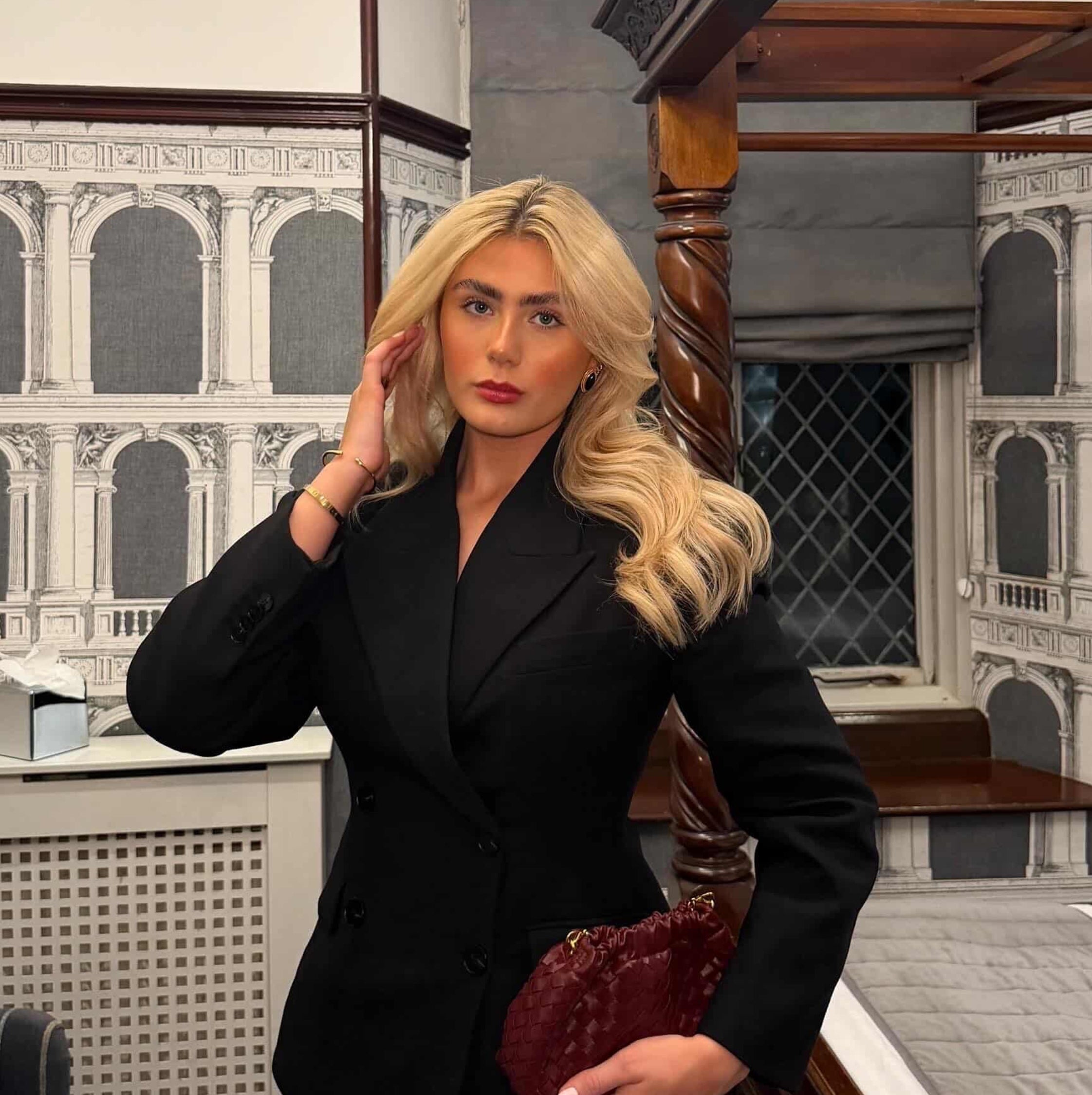 Woman in a black suit standing in an elegant interior setting with chandelier and staircase.