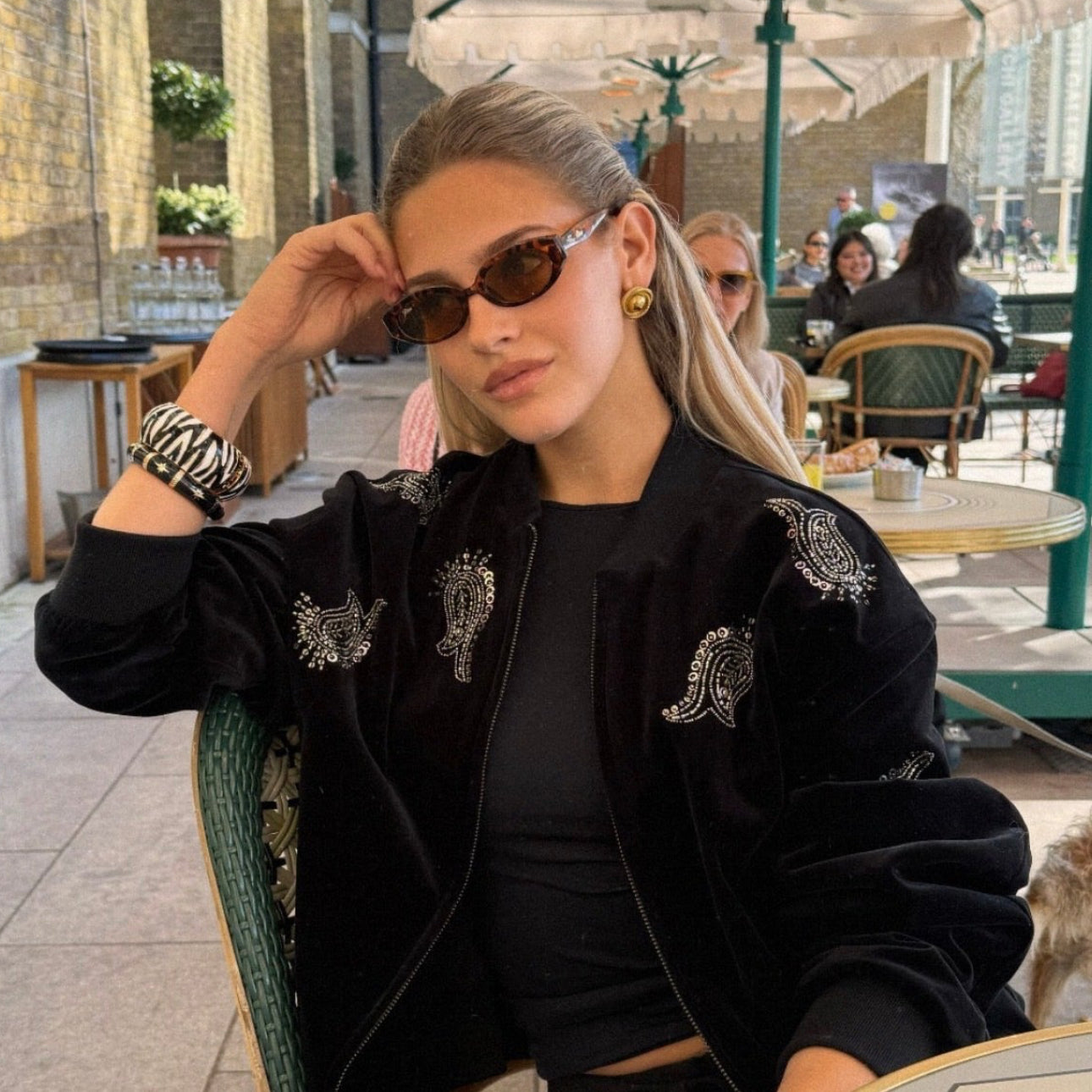 Woman sitting outdoors in a casual setting wearing sunglasses and a black jacket.